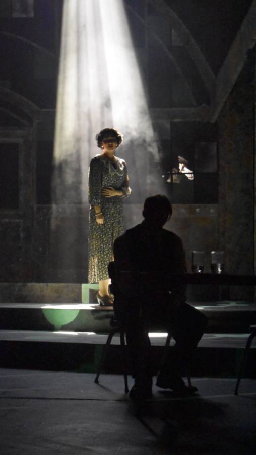 Image of lady standing in dark room with a ray of sunlight shining down on her while the sillouett of a man is sitting in front of her facing away, head down